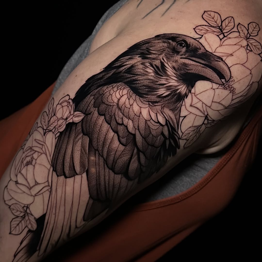 Detailed black and grey raven with spread wings surrounded by delicate flowers on upper arm, featuring realistic shading and fine linework.