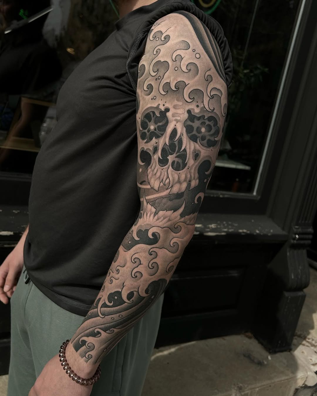 Full arm sleeve tattoo featuring an ornate skull with scrollwork patterns in black and grey shading on a person's upper arm and forearm.