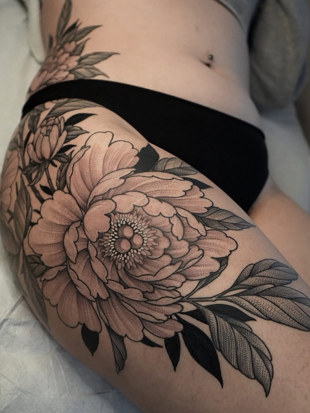 Large black and grey peony flower tattoo on upper arm with detailed shading, leaves, and fine line work.