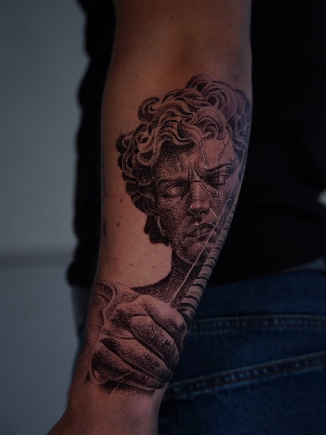Classical male bust with curly hair and ornate staff on forearm, rendered in black and grey with smooth shading gradients.