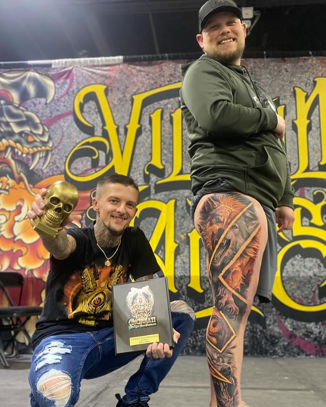 Large black and grey demon or oni face with flowing hair and shading on male's leg, displayed at tattoo event.