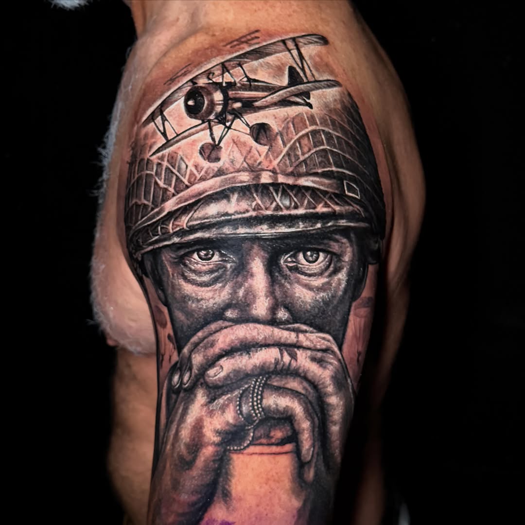 Black and grey portrait tattoo of a bearded man wearing a winged helmet with a biplane flying above his head on the upper arm.