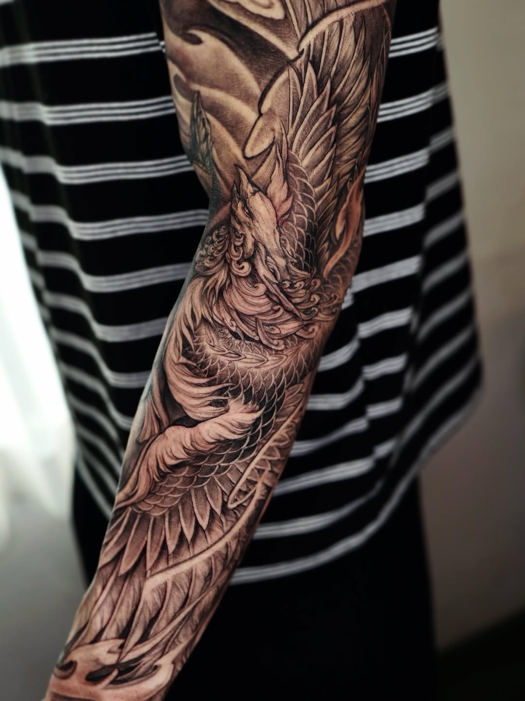 Detailed phoenix wing tattoo on forearm with intricate feather texturing and smooth black and grey shading, worn by person in striped shirt.